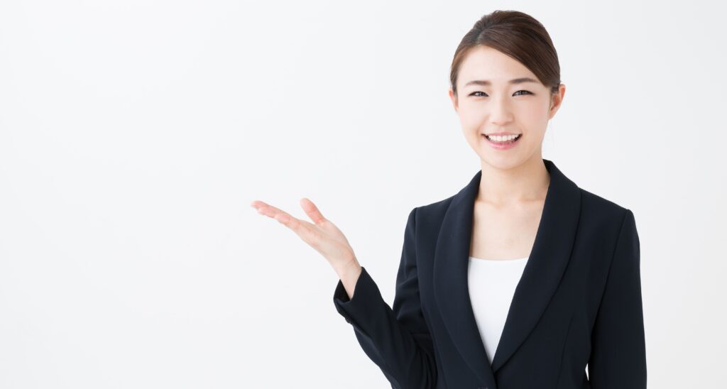 黒のスーツを着た笑顔の女性が、右手を上に上げ、紹介するかのように差し出している様子