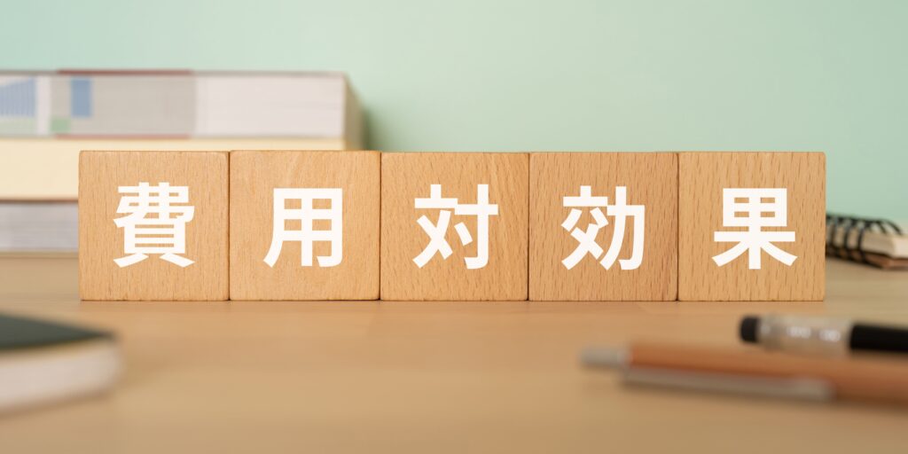 デスク上に置かれた木製ブロックに、白い文字で「費用対効果」と書かれているクローズアップ。ビジネスや効率をテーマにしたイメージ。